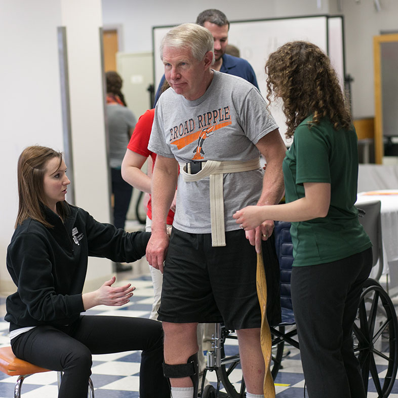 Two physical therapy students helping a wheelchair bound guy walk.